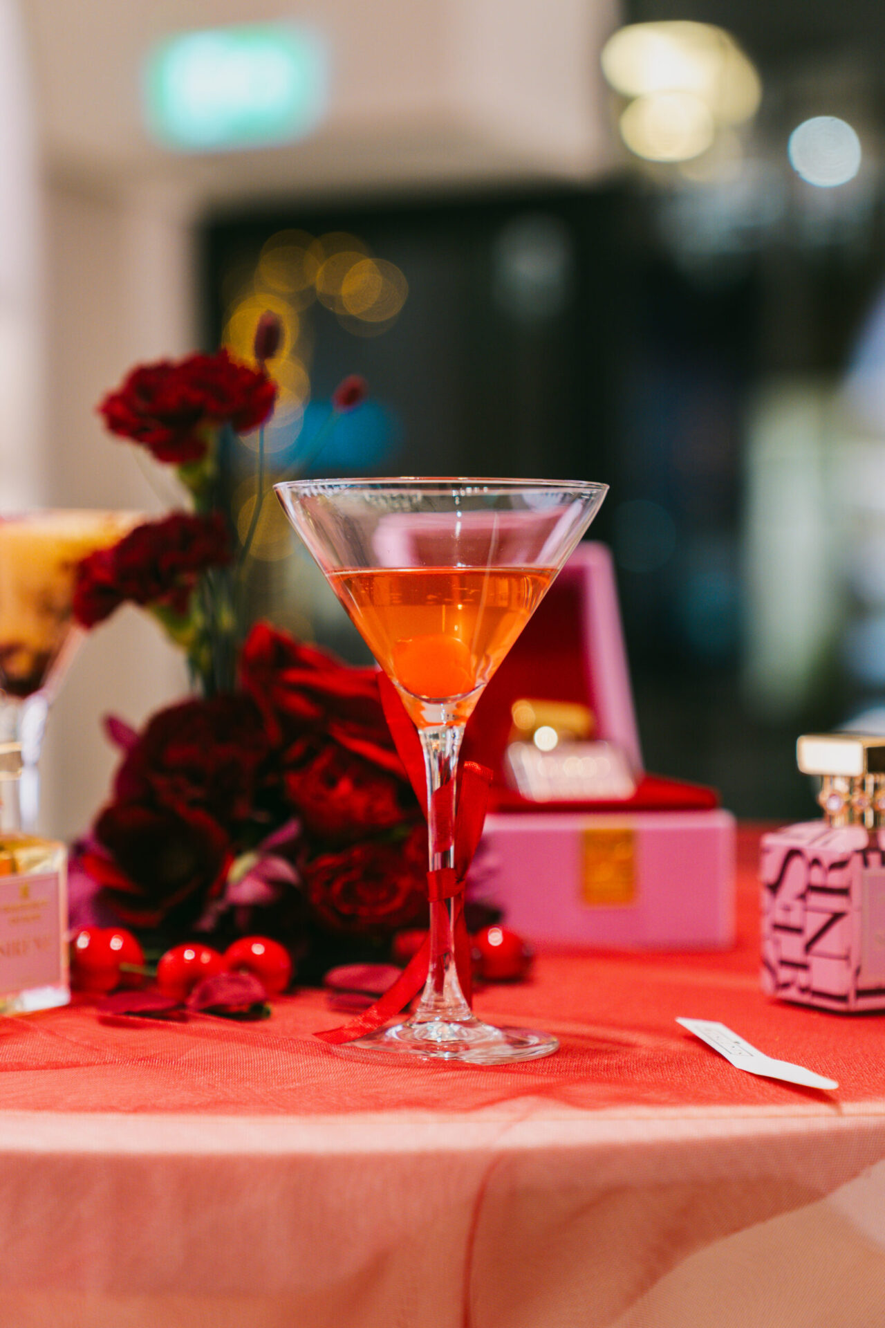 Event styling Een cocktail in een martini-glas met een oranje drank, staat op een tafel met een rode tafelkleed. Er zijn ook rode rozen en parfumflessen zichtbaar, passend bij de Wescent niche parfum lancering.